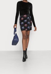 Kvinna klädd i en svart långärmad topp och en svart minikjol med paljettryck av läppar, hållande en marinblå Valentino-väska och bärande svarta ankelskor.