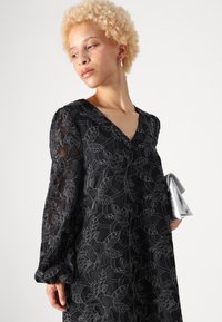 Fekete ruha virágos hímzéssel, V nyakkivágással, hosszú ujjakkal, texturált anyagból, ezüst clutch-csal párosítva, réteges mintákat bemutatva.