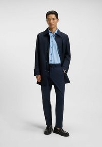 Cappotto lungo blu navale sopra una camicia a righe azzurre chiare e pantaloni coordinati. Scarpe nere senza lacci completano l'outfit. Design minimalista, vestibilità su misura.
