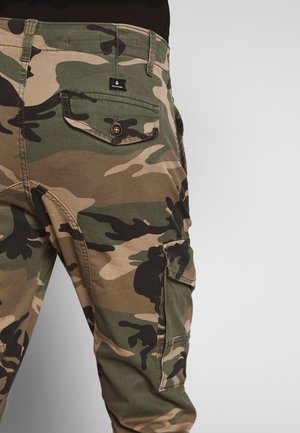 Onderlichaam draagt groene en bruine camouflerende cargobroek met met knopen afgesloten achter- en zijzakken op een neutrale achtergrond.