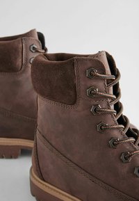 Botas de senderismo de cuero marrón con soporte de ante en el tobillo, ojales de metal y cordones texturizados. Suelas de goma estriadas. Detalle de costura sutil.