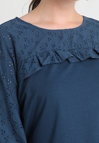 Blusa blu con una sovrapposizione in pizzo con ritagli floreali e un accento arricciato. Il tessuto è morbido con una superficie liscia.
