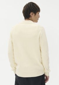 Krém színű kötött pulóver bordázott textúrával, gallérral és hosszú ujjakkal, hátulról bemutatva. Testhez simuló fazon egyenes szegéllyel.