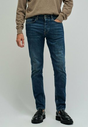 Hombre vistiendo jeans ajustados azul oscuro, suéter marrón y mocasines de cuero negro, de pie con una mano en el bolsillo sobre un fondo gris.