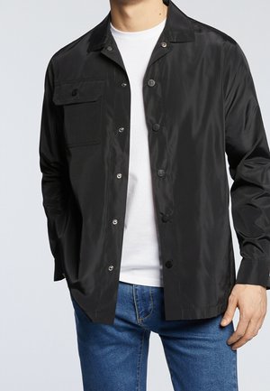 Homme portant une veste noire boutonnée sur un t-shirt blanc et un jean bleu, les mains détendues le long du corps, devant un fond uni.
