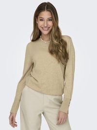 Pull en tricot beige à manches longues, avec des poignets côtelés et des accents de boutons. Associé à un pantalon de couleur claire, texture lisse.