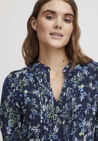 Temno modra bluzica s cvetličnim vzorcem v zeleni in beli barvi, z V-izrezom, nabranim oblikovanjem in mehko, lahko tkanino.