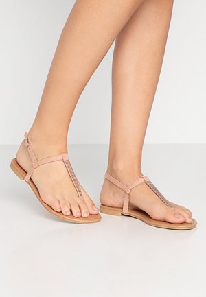 Pies con sandalias planas de color rosa claro con tira en T fina y hebilla pequeña, de pie sobre una superficie lisa y clara.