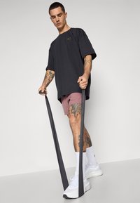 Crna oversized majica s logotipom, ružičaste kratke hlače i bijele tenisice. Model drži crne otporne trake s vidljivim tetovažama na rukama.