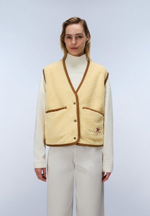Kvinde iført en cremefarvet rullekrave og lys beige bukser med en ærmeløs beige fleecevest med brun kant og forlommer.