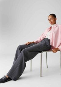Lichtroze overhemd met knopen, grijze wijde broek met aantrekkoord in de taille, zwarte loafers, zittend op een metalen stoel tegen een effen achtergrond.
