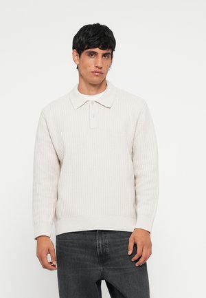 Light beige knit pullover with a collared neckline, textured surface, long sleeves, and ribbed cuffs, paired with dark grey jeans.