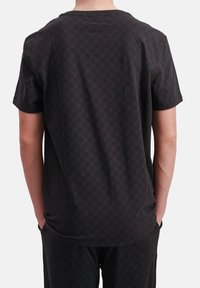 Persoon in een zwart T-shirt met korte mouwen en bijpassende broek, staand met de handen in de zakken, gezien van achteren.