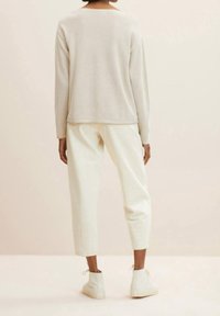 Persoon die een beige trui draagt, off-white cropped broek en witte hooggesloten schoenen, gezien van achteren tegen een effen lichte achtergrond.