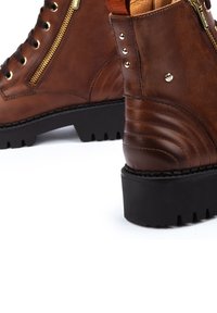 Bottines en cuir marron avec lacets, fermeture éclair sur le côté, bout arrondi, panneaux texturés, quincaillerie dorée, et épaisse semelle en caoutchouc noir.