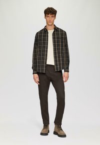 Karierter Button-up-Hemd in Schwarz und Beige, über einem cremefarbenen Pullover, kombiniert mit dunkelbraunen Hosen und braunen Schnürstiefeln.
