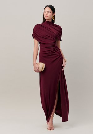 Vrouw draagt een vloerlange bordeauxrode jurk met geplooide details, houdt een beige clutch vast en is voorzien van gouden oorbellen en armband.