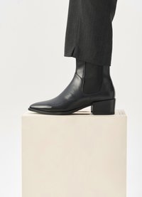 Μαύρο δερμάτινο μποτάκι μέχρι τον αστράγαλο με μυτερή μύτη, ελαστική πλευρική πάνελ και χαμηλό τετράγωνο τακούνι, εκτεθειμένο σε μια ανοιχτή βάση σε ουδέτερο φόντο.
