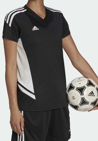 Camiseta atlética negra con acentos blancos y tres franjas blancas en los hombros; combinada con pantalones cortos negros. Se sostiene un balón de fútbol con una mano.