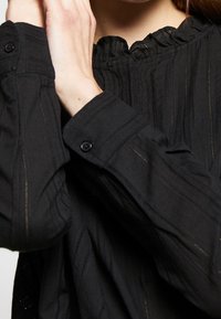 Svart, texturerad blus med en volangkrage, vertikal randig mönstring och knappstängningar på ärmarna.