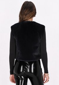 Vest din blană sintetică navy cu un design sleek și scurt, purtat peste un top negru cu mânecă lungă, asociat cu colanți negri strălucitori.