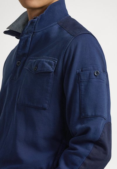 Mornarsko moder pulover z visokim ovratnikom, dvema prsnikoma z žepi, zapenjanjem na gumbe in kontrastnimi ramenimi paneli. Gladek tekstura, sproščen dizajn.