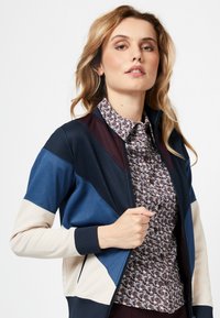 Multikleurige jas met marineblauwe, beige en bordeauxpanelen, met een rits aan de voorkant en ribgebreide manchetten, gedragen over een gemusterd shirt.