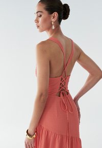 Koronkowa sukienka w kolorze koralowym z zapięciem na plecach, cienkimi ramiączkami i rozkloszowanym dołem. Posiada związany detal w talii oraz gładkie, czyste wykończenie.