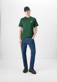 T-shirt en coton vert avec logo, jeans en denim bleu, baskets noires et casquette noire. Le modèle se tient devant un fond blanc.