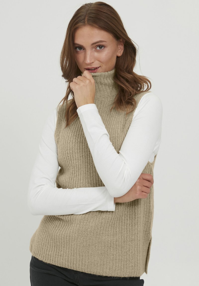 Pull à col montant sans manches en maille côtelée beige, porté sur une chemise blanche à manches longues. Texture douce et coupe décontractée.