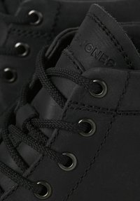 Zapatos de cuero negro con una textura suave, cordones negros y ojetes de metal. El nombre de la marca "Jones" está grabado en el lado.