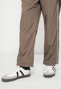 Λευκά αθλητικά παπούτσια με μαύρες ρίγες και γκρίζες λεπτομέρειες, συνδυασμένα με χαλαρούς καφέ παντελόνες που διαθέτουν λευκή ρίγα στο πλάι και λεία υφή.