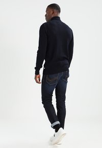 Marineblå strikkegenser med et strukturert fiskebeinmønster, kombinert med mørkeblå slim-fit jeans og hvite sneakers, med ryggen mot betrakteren.