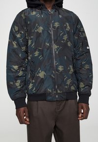 Kurtka bomber w kamuflażu w ciemnozielonym i czarnym kolorze, z zamkiem błyskawicznym z przodu, ściągaczami przy mankietach oraz wyściełanym kapturem dla dodatkowego ciepła.