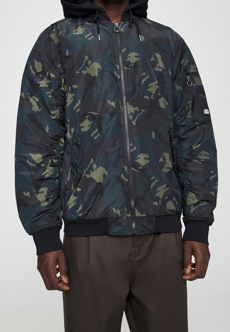 Kurtka bomber w kamuflażu w ciemnozielonym i czarnym kolorze, z zamkiem błyskawicznym z przodu, ściągaczami przy mankietach oraz wyściełanym kapturem dla dodatkowego ciepła.