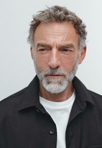 Ritratto di un uomo con capelli ricci grigi e barba, che indossa una camicia nera a bottoni sopra una maglietta bianca, con espressione seria.