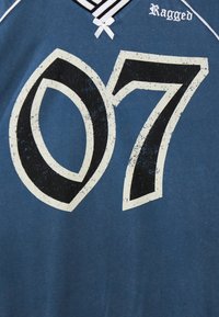 Modrá tričko s veľkým, opotrebovaným čiernym číslom "07" s bielym obrysom. Golier má čierno-biely pruhovaný vzor a biely mašľový akcent.