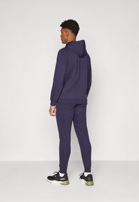 Marineblauwe hoodie en bijpassende joggers van zacht materiaal. Voorzien van ribbelmanchetten, een ontspannen pasvorm en zwarte sportschoenen.
