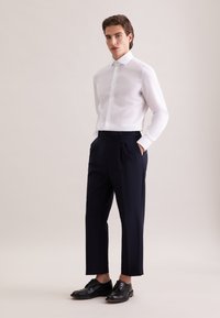 Witte overhemd met knopen, donkerblauwe geplooide broek en zwarte leren schoenen. Het overhemd is glad; de broek heeft een getailleerde pasvorm en de schoenen hebben veters.