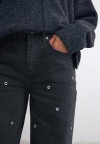 Schwarze Jeans aus Denim mit Ösen-Details, im High-Waist-Stil und mit vorderen Taschen. Kombiniert mit einem dunkel strukturierten Pullover.