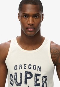 Witte katoenen tanktop met marineblauwe bedrukte tekst "OREGON SUPER." Heeft een ronde halslijn en brede schouderbanden. Eenvoudig ontwerp.