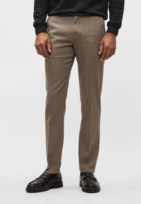 Vaaleanruskeat räätälöidyt housut, joissa on sileä kangaspinta, suora leikkaus edestä ja sivutaskut, yhdistettynä mustiin loafereihin.