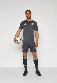 Grijs voetbaltenue met korte mouwen en gouden accenten. Bevat het "OL"-logo, gecombineerd met zwarte korte broek en sokken met witte strepen. Houdt een zwart-witte voetbal vast.