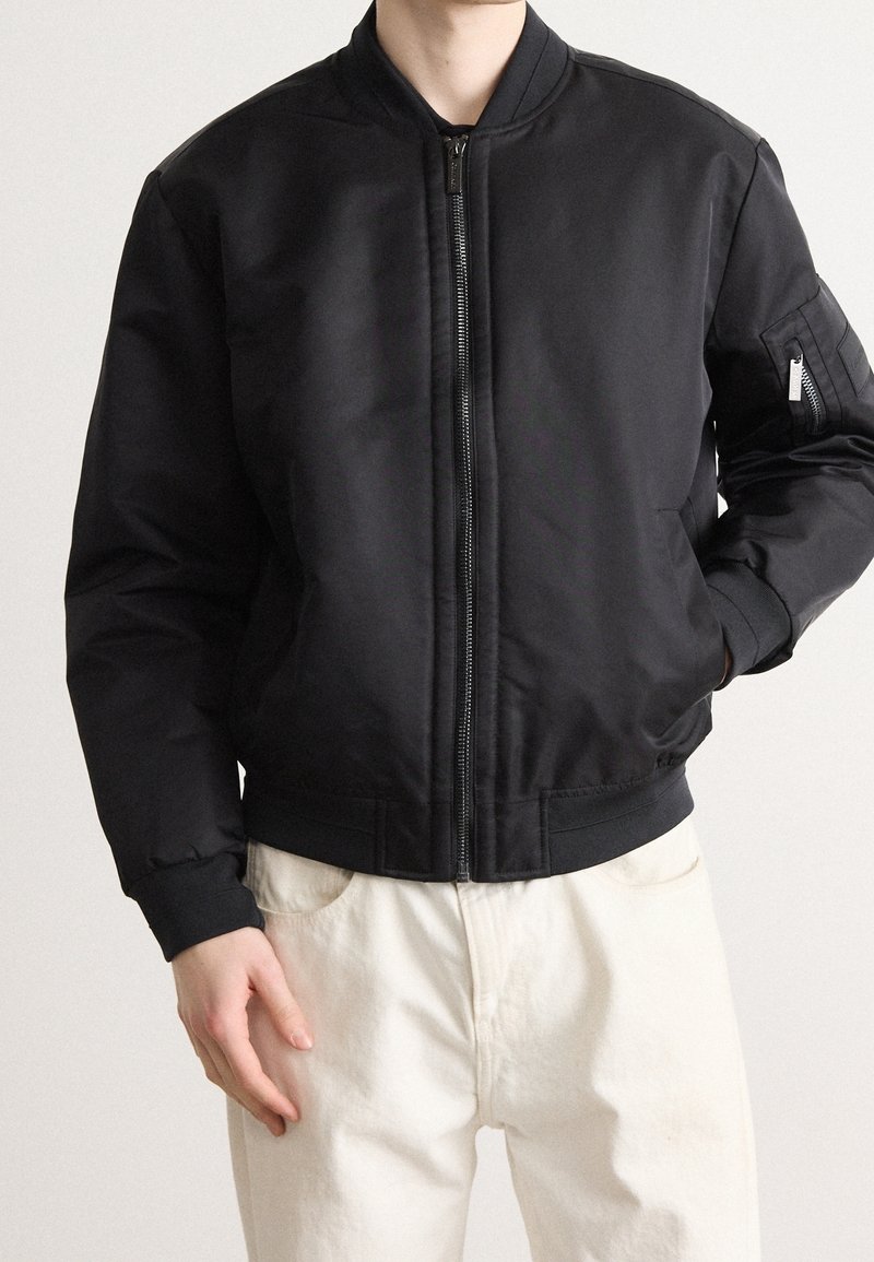 Schwarze Bomberjacke mit Reißverschluss, gerippten Bündchen und Saum. Glattes, synthetisches Material. Verfügt über Seitentaschen und eine Akzenttasche am Arm.