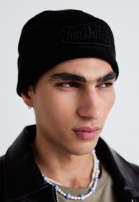 Gorro negro con el logo en relieve "Von Dutch Originals", textura de punto, llevado por un modelo con una chaqueta de cuero y collares en capas.