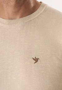 Lichtbeige gebreide trui met een ronde hals. Bevat een klein bruin geborduurd vogellogo aan de bovenlinkerzijde. Zachte textuur.