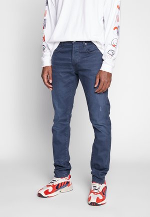 Man wearing dark blue slim-fit jeans paired with white and red athletic sneakers and a white long-sleeve shirt with graphic sleeves.