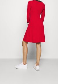 Punainen ribbineulemekko, jossa on pitkät hihat, leveähelma ja kontrastina mustavalkoiset raidalliset yksityiskohdat kaula-aukkossa ja hihoissa. Yhdistetty valkoisiin tennareihin.