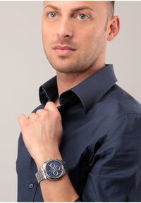 Silverfärgad rostfri klocka med en blå urtavla, tre subdials och upplysta visare. Buren på ett handled, matchad med en m örkblå skjorta.