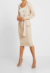 Cardigan beige côtelé avec boutons, taille cintrée ; associé à une jupe midi assortie. Porté avec des sandales à talons hauts à lanières métalliques.